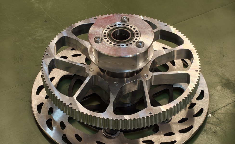 The driven-side wheel hub assembly — a single machined-aluminium part combining the belt's final-drive pulley, the wheel hub bearing, and the brake rotor mount