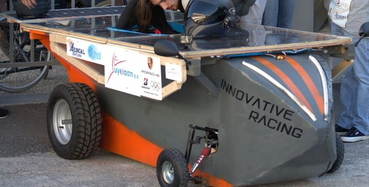 The Innovative Racing solar car in side view on race day, its grey and orange bodyshell under a flat roof deck of solar cells