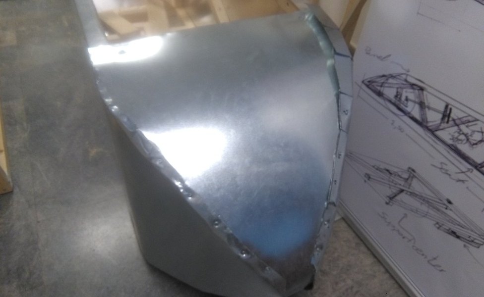 The car&rsquo;s aluminium nose section during fabrication, standing on a workshop floor: a curved sheet-metal skin riveted along a folded flange, with a whiteboard of hand-drawn frame and suspension sketches propped behind it