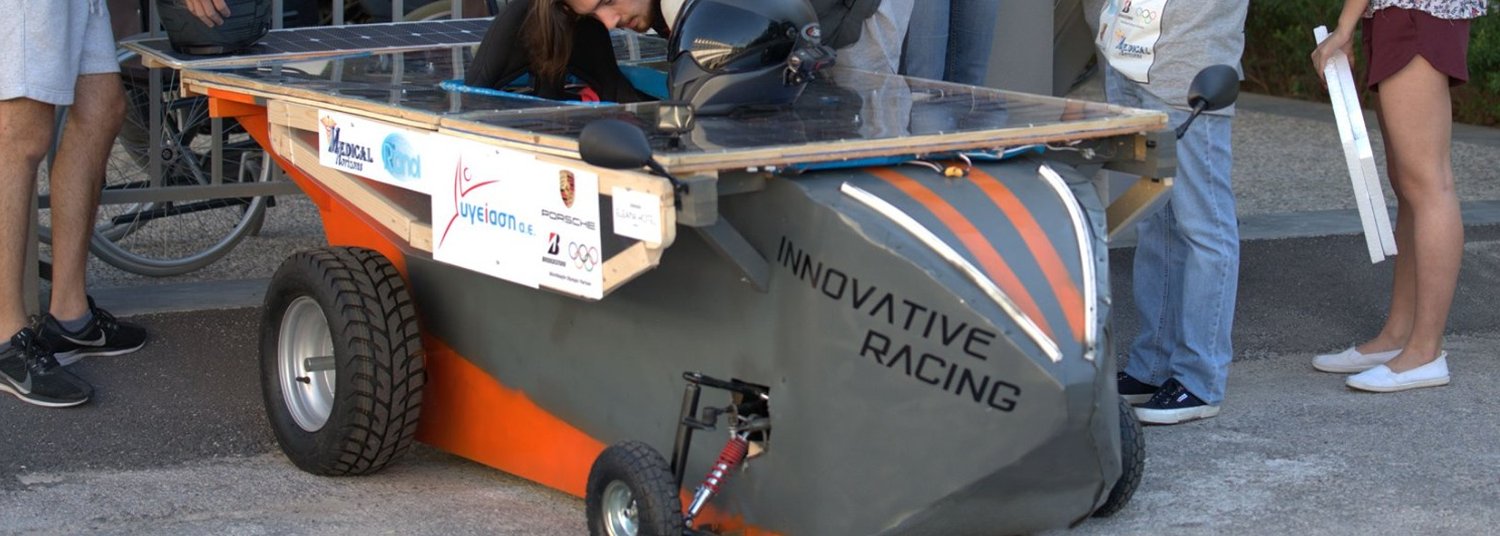 The Innovative Racing solar car at the race, seen from the side: a grey and orange bodyshell on a wooden frame, a flat roof deck of solar cells above it, knobbly kart tyres, and an exposed coil-over damper at the rear wheel
